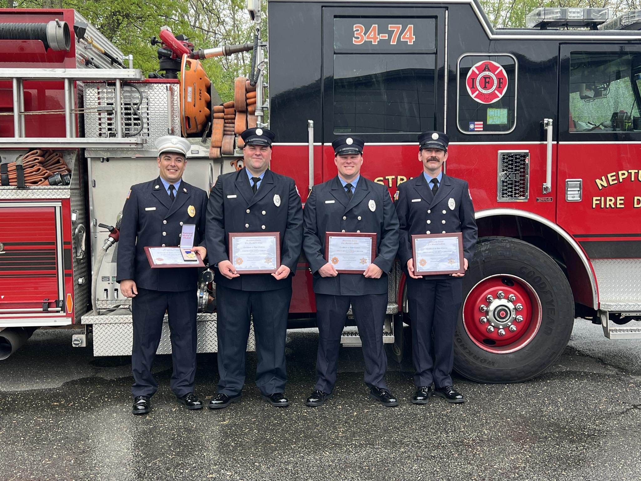 Four Neptune Township firefighters stand in dress uniforms with awards in front of a fire engine