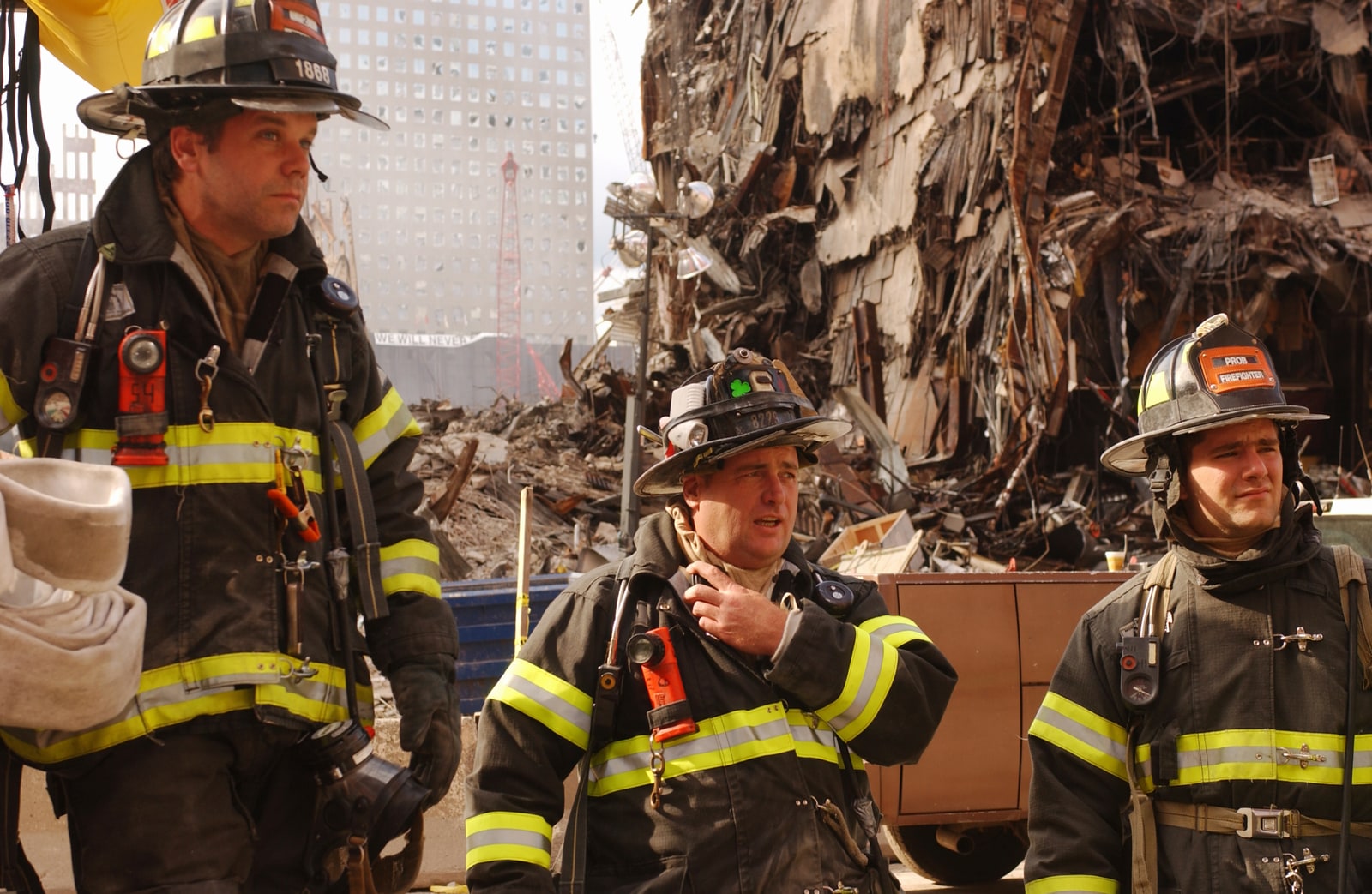 Three firefighters in turnout gear stand amid damaged buildings and debris at the World Trade Center site.