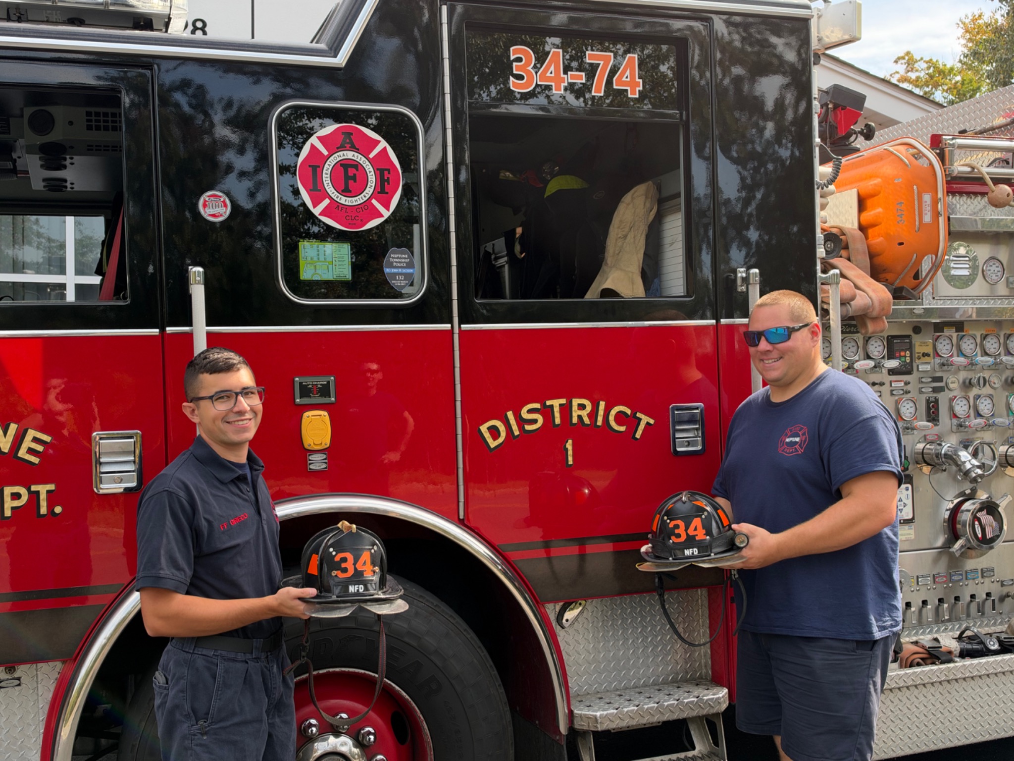 Firefighters Michael Greco and Frank Martuscelli Jr. holding their helmets beside a Neptune fire engine.